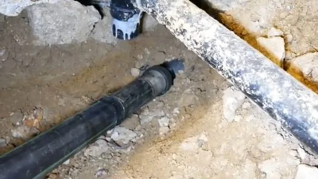 A length of black drain pipe laid in a trench cut through a broken concrete slab with a second pipe running alongside