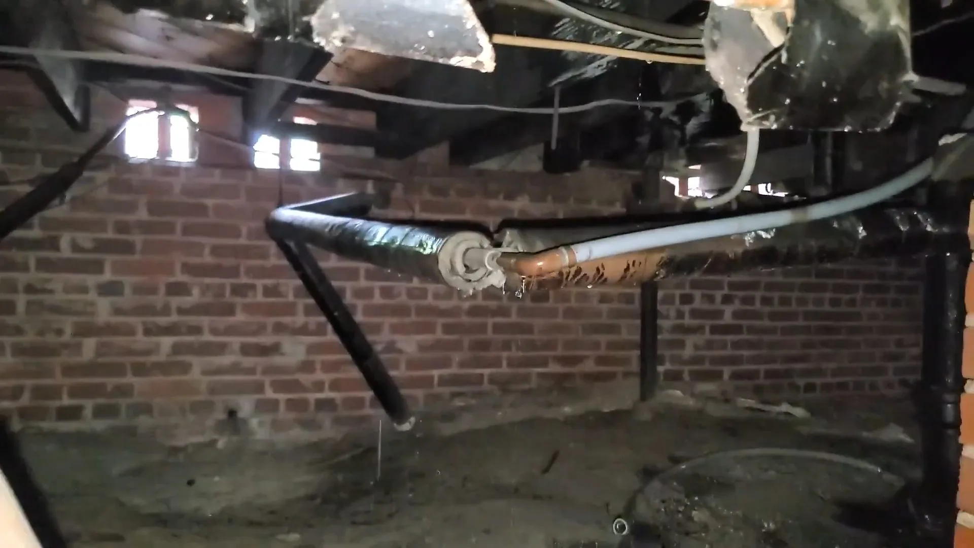 A run of black steel pipe with a valve and white tubing along the joists of a brick-walled crawl space