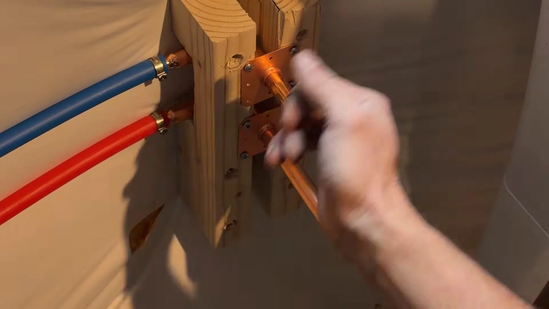 A hand adjusting a copper stub-out fixed to a wooden backer, with blue and red PEX lines connected on the left