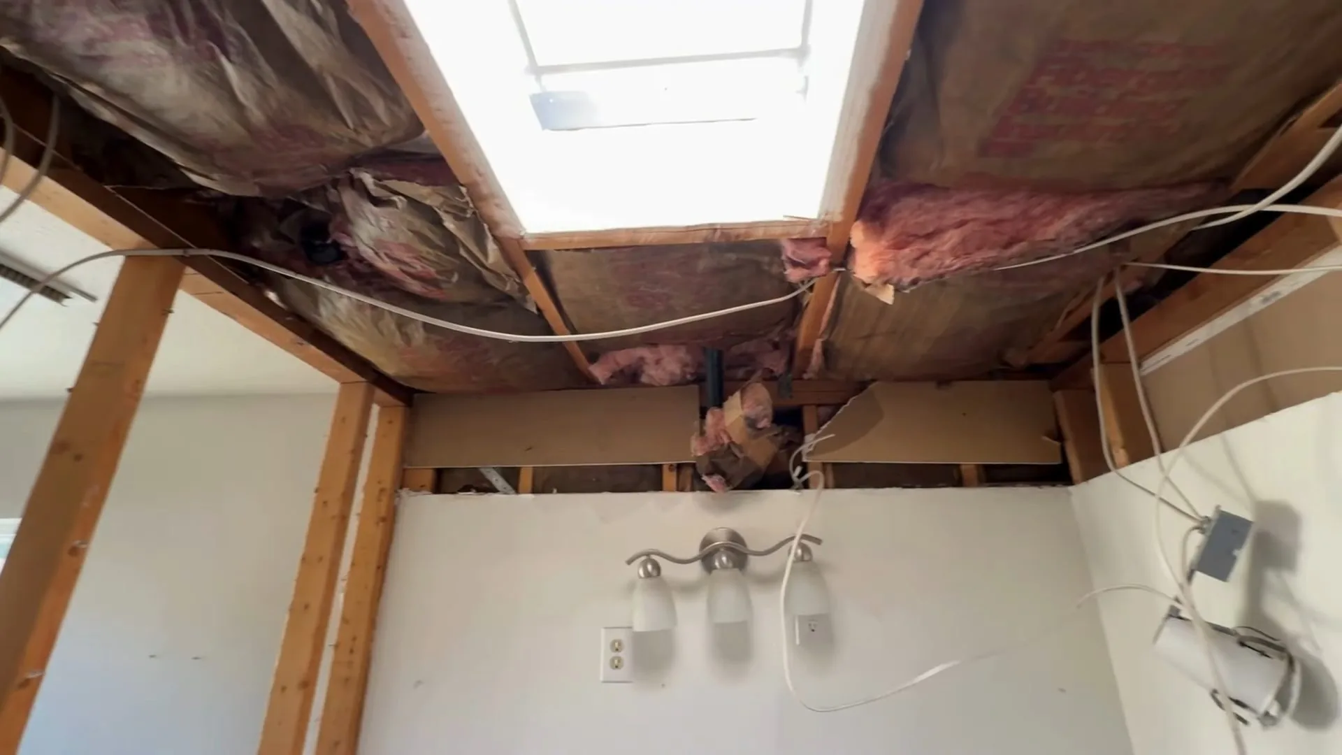 A bathroom ceiling opened up to expose pink insulation, framing and wiring above a bathtub