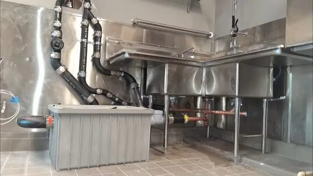 A commercial kitchen with stainless steel sinks, exposed drain lines and a grease interceptor tank on the tile floor