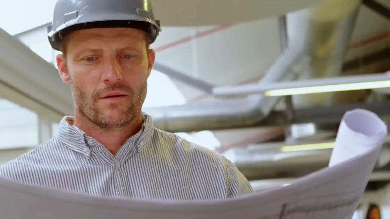 A man in a hard hat looking down at a set of rolled plans inside an unfinished building