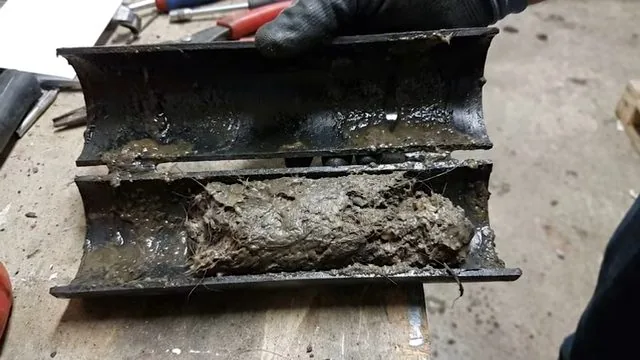 A length of black drain pipe cut open lengthwise to show a thick grey deposit packed along the bottom
