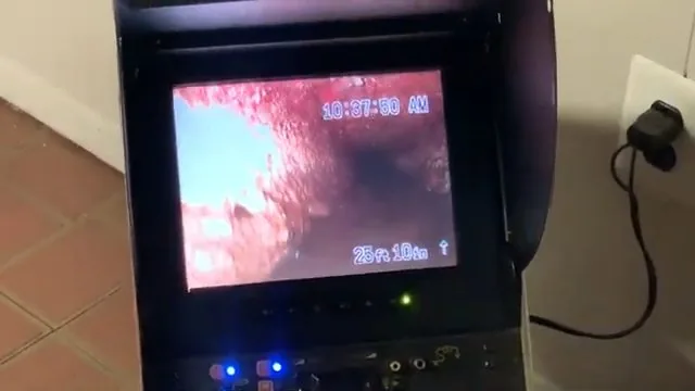 A drain inspection monitor on a tiled floor showing the reddish inside of a pipe, with a timestamp on screen