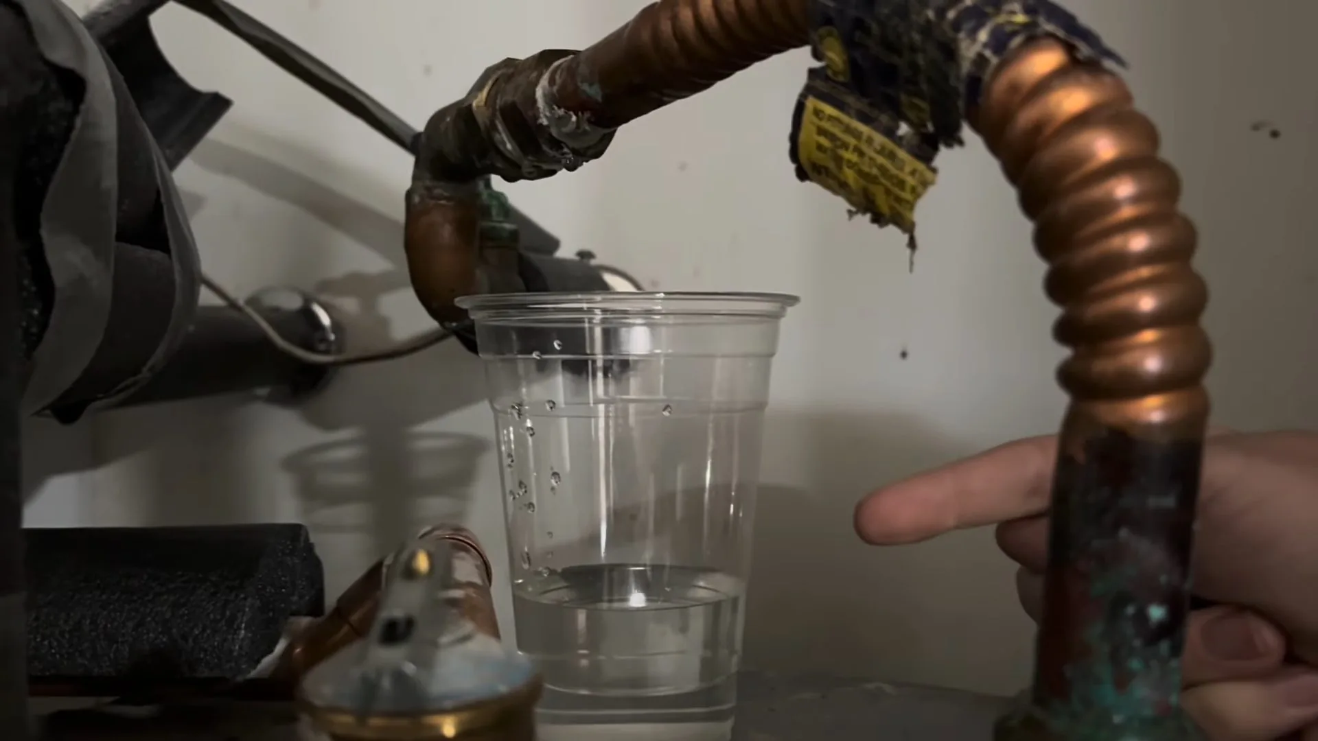 A hand opening a drain valve on a water heater while a clear cup catches the water, with copper connectors above