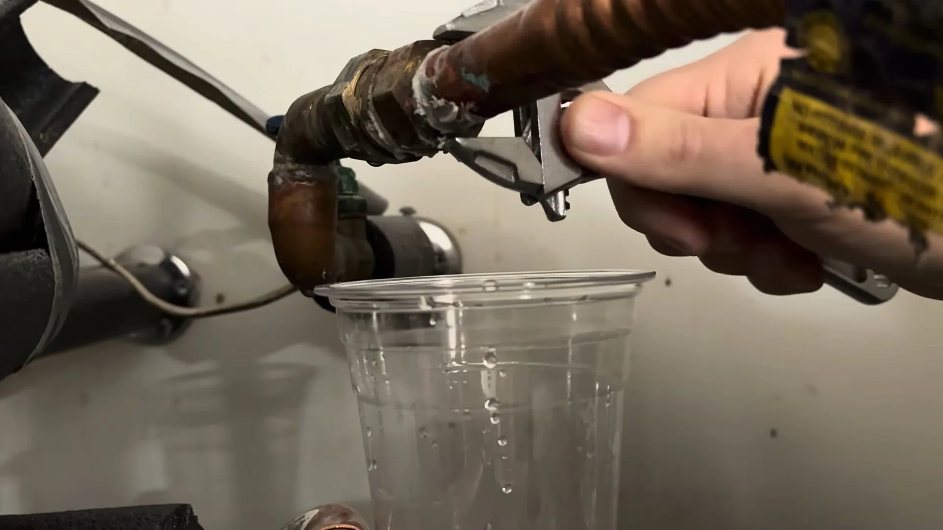 A technician holding a wrench on a water heater drain connection while water runs into a clear plastic cup