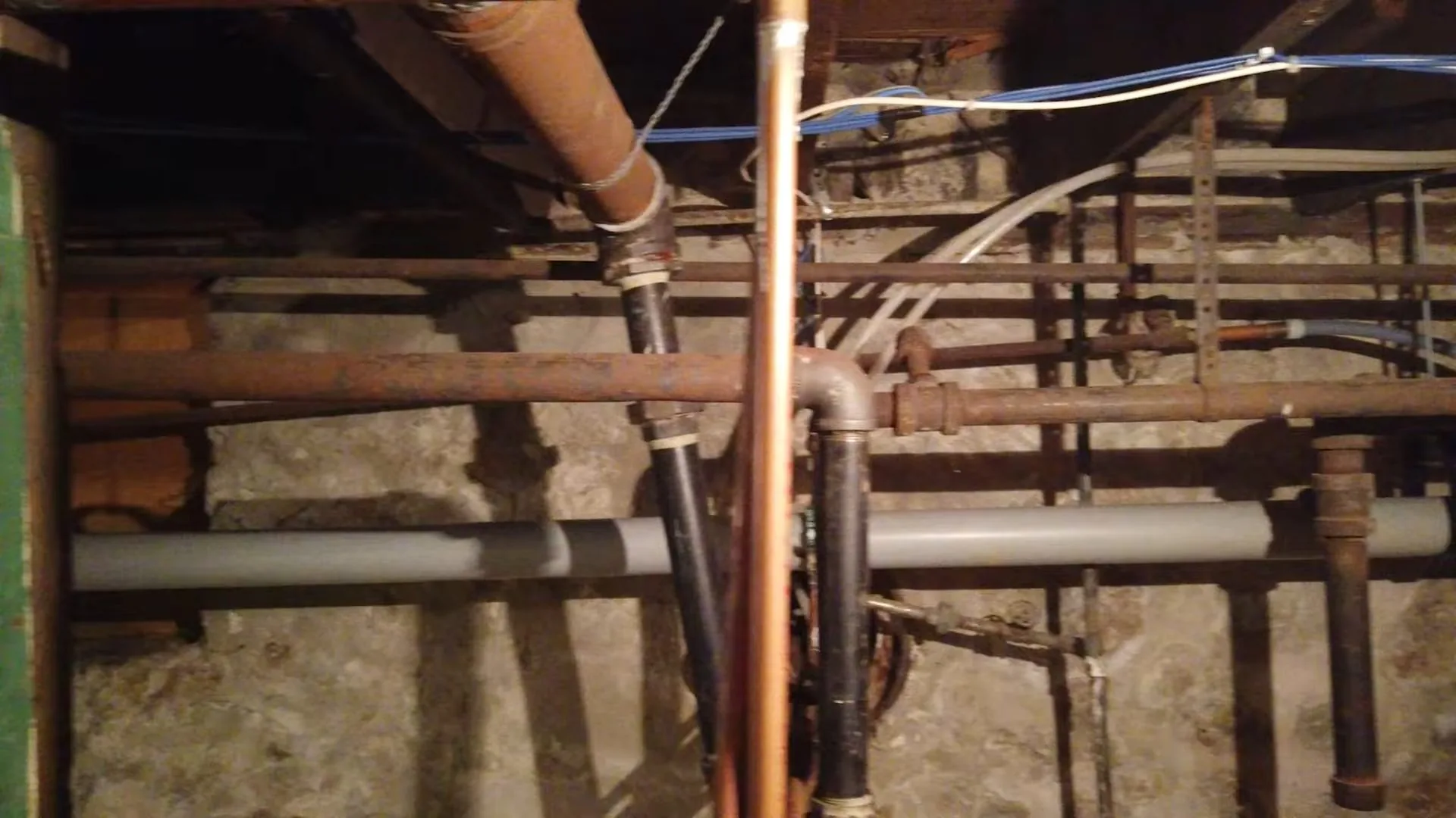 A basement ceiling with copper, iron and plastic pipes running together between wooden joists