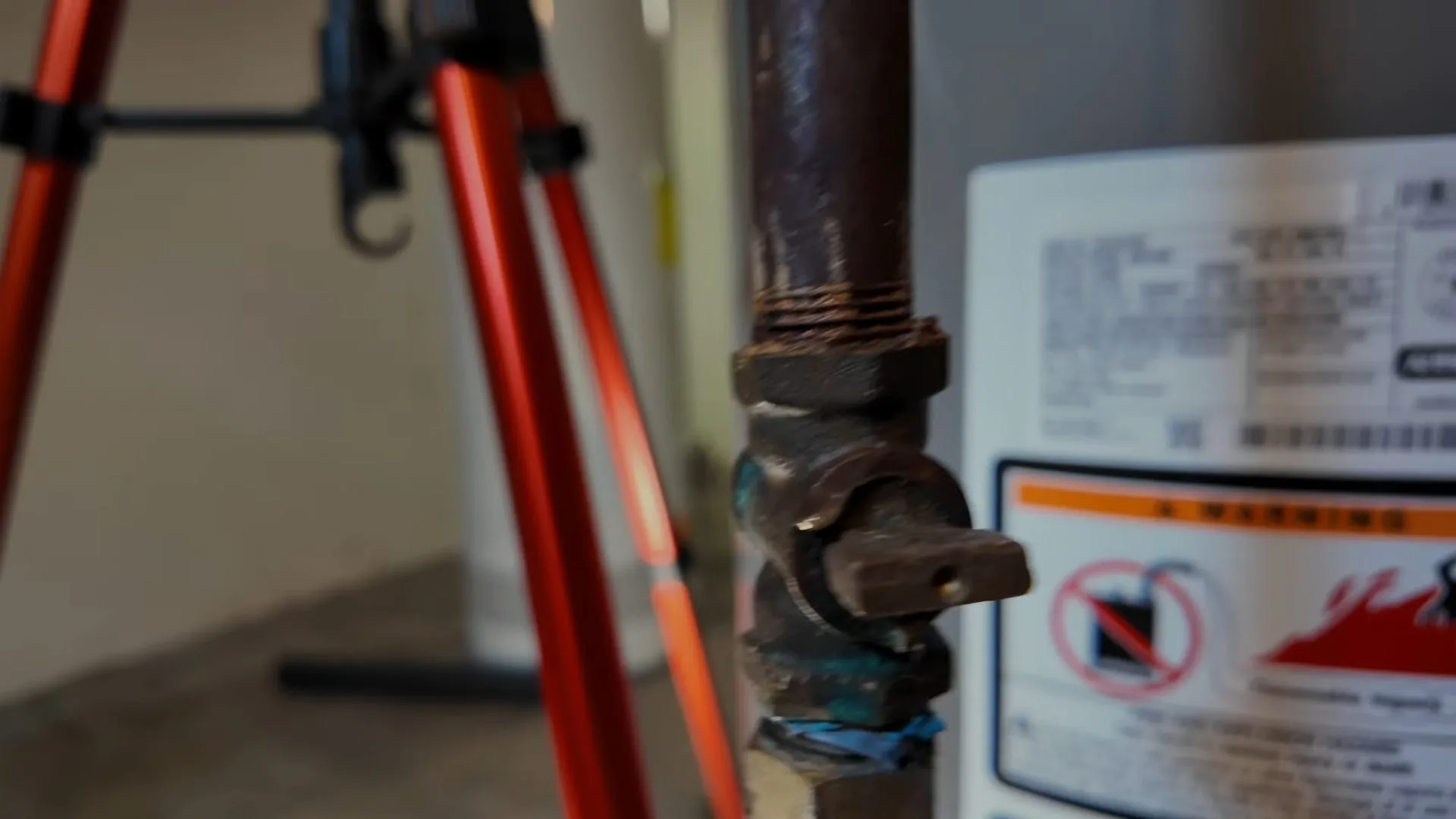 A black iron gas shutoff valve with a stuck blue-green fitting beside a water heater, with an orange tripod behind