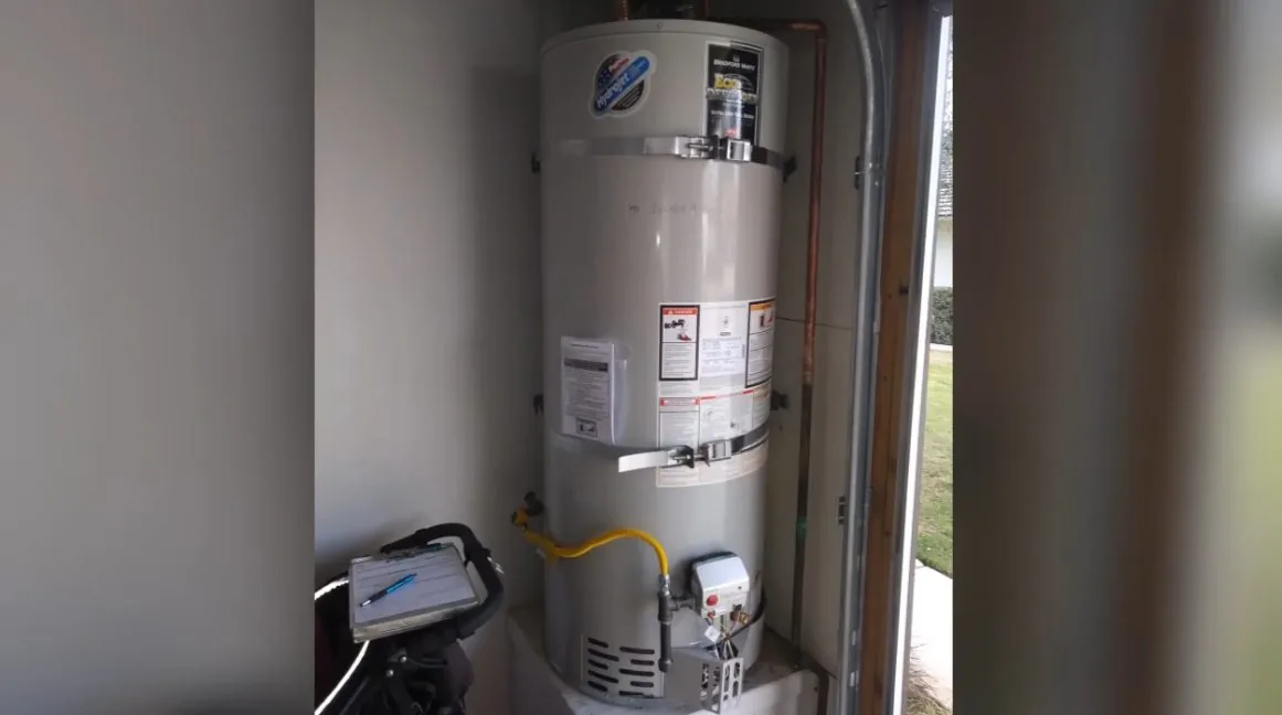 A tall gas tank water heater standing in a utility closet with a yellow gas connector and a small device on the floor