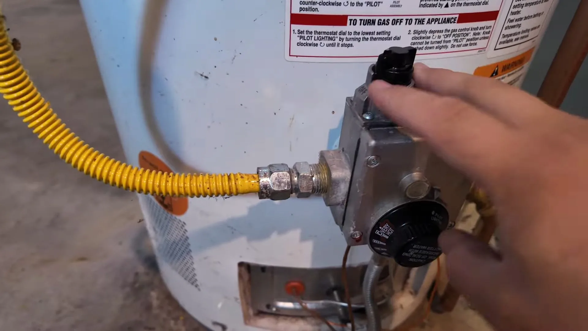 Hand turning the control dial on a gas water heater thermostat beside a yellow flexible gas connector