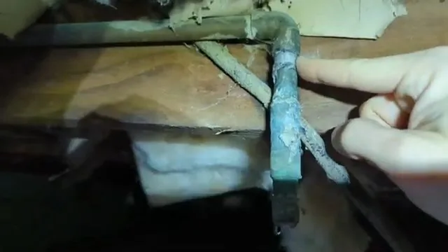 A fingertip resting on a copper pipe corner coated in green corrosion under a floor with insulation