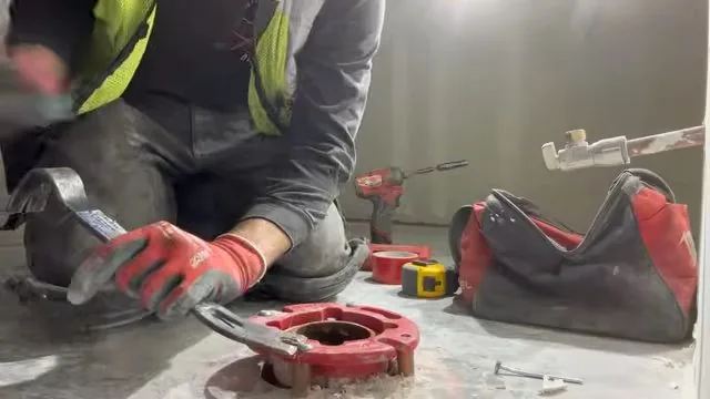 A worker in a yellow vest kneeling beside a red toilet flange ring and a tool bag on a concrete floor