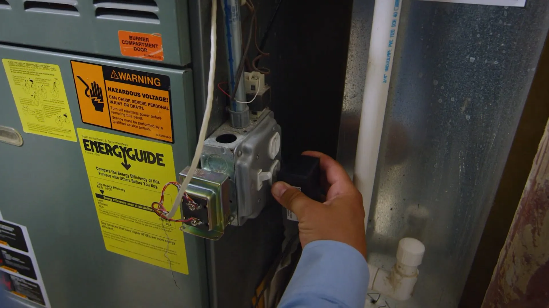 A hand on the gas valve of a furnace with warning and energy labels on the cabinet
