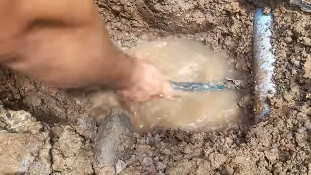 A hand reaching into a muddy, water-filled excavation beside a pale pipe stub