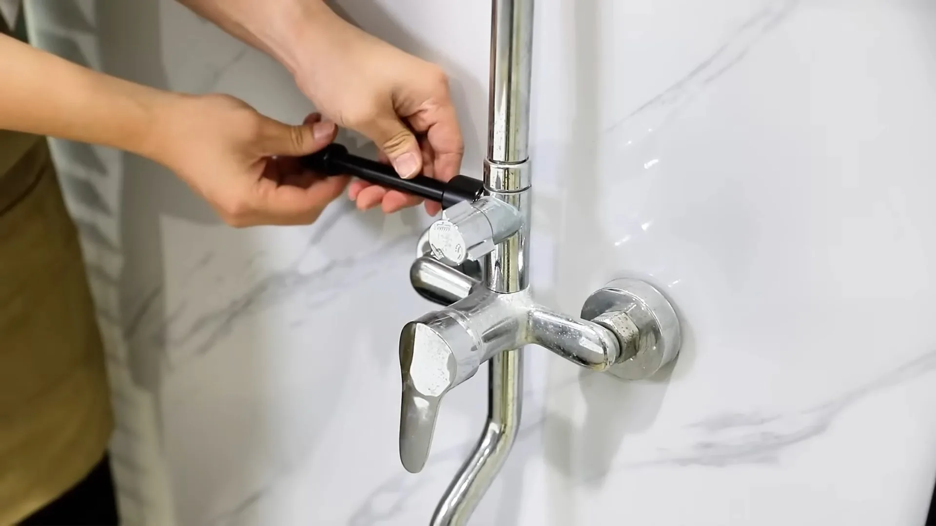 Two hands using a small tool on a chrome wall-mounted tub and shower faucet set against white marble