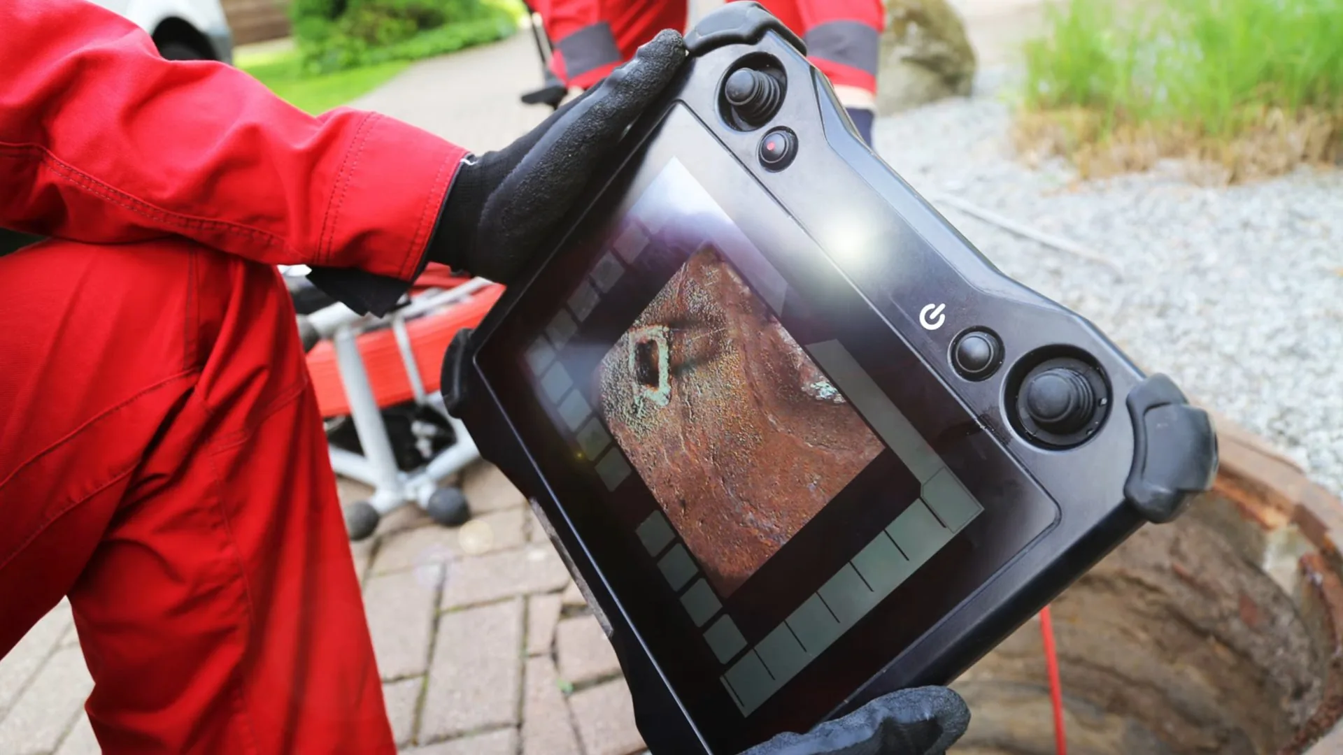 A person in a red jacket holding a handheld monitor that shows the inside of a pipe during an inspection