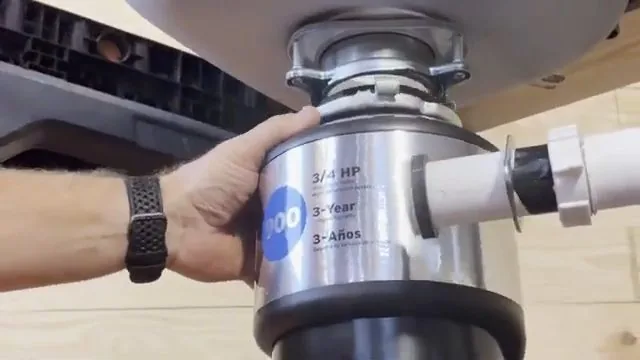 A pair of hands holding a stainless garbage disposal in place under a kitchen sink with a drain pipe attached