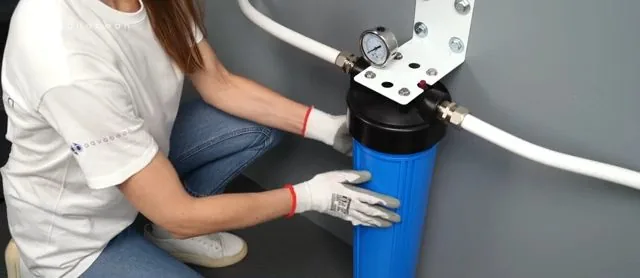 A person kneeling to fit a blue filter housing on a wall bracket with white supply pipe on either side
