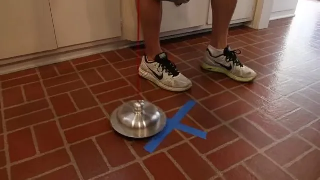 A metal listening disc set on a terracotta tile floor next to a blue tape cross, with a person standing close by