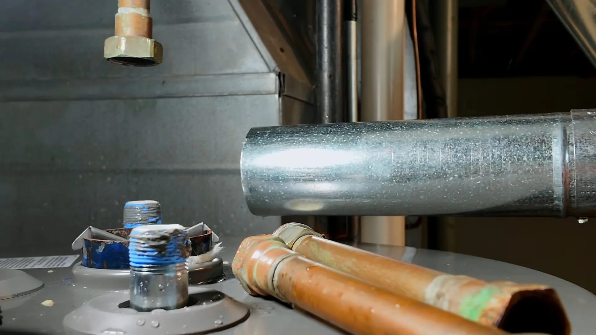 The top of a water heater tank with copper pipe stubs, braided connectors and a vent pipe beside it