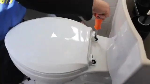 A white toilet with a hand and an orange-handled tool working on the base bolt cap