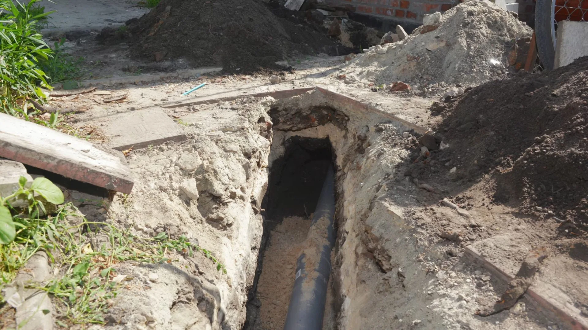 A narrow trench dug beside a house with a dark sewer pipe visible at the bottom and soil piled on both sides
