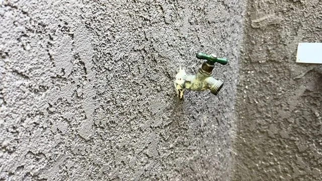 A brass outdoor hose bib with a green handle on a grey stucco wall
