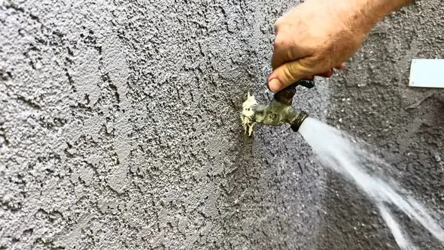 A brass hose bib on a stucco wall with a strong stream of water running from the spout