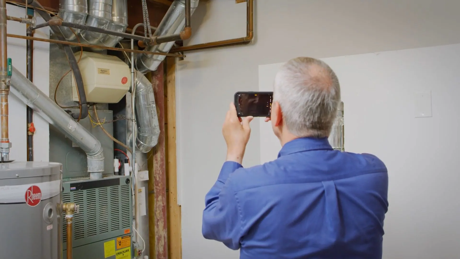 A man in a blue shirt holding a phone up to photograph a water heater and furnace area