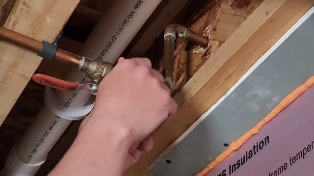 A hand holding a copper and PEX connection with a red valve handle inside an open wall cavity