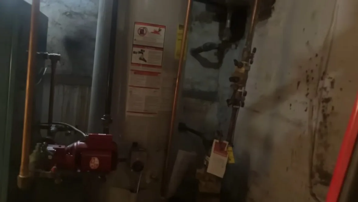 An older gas water heater with a red circulating pump and exposed copper pipe in a utility room
