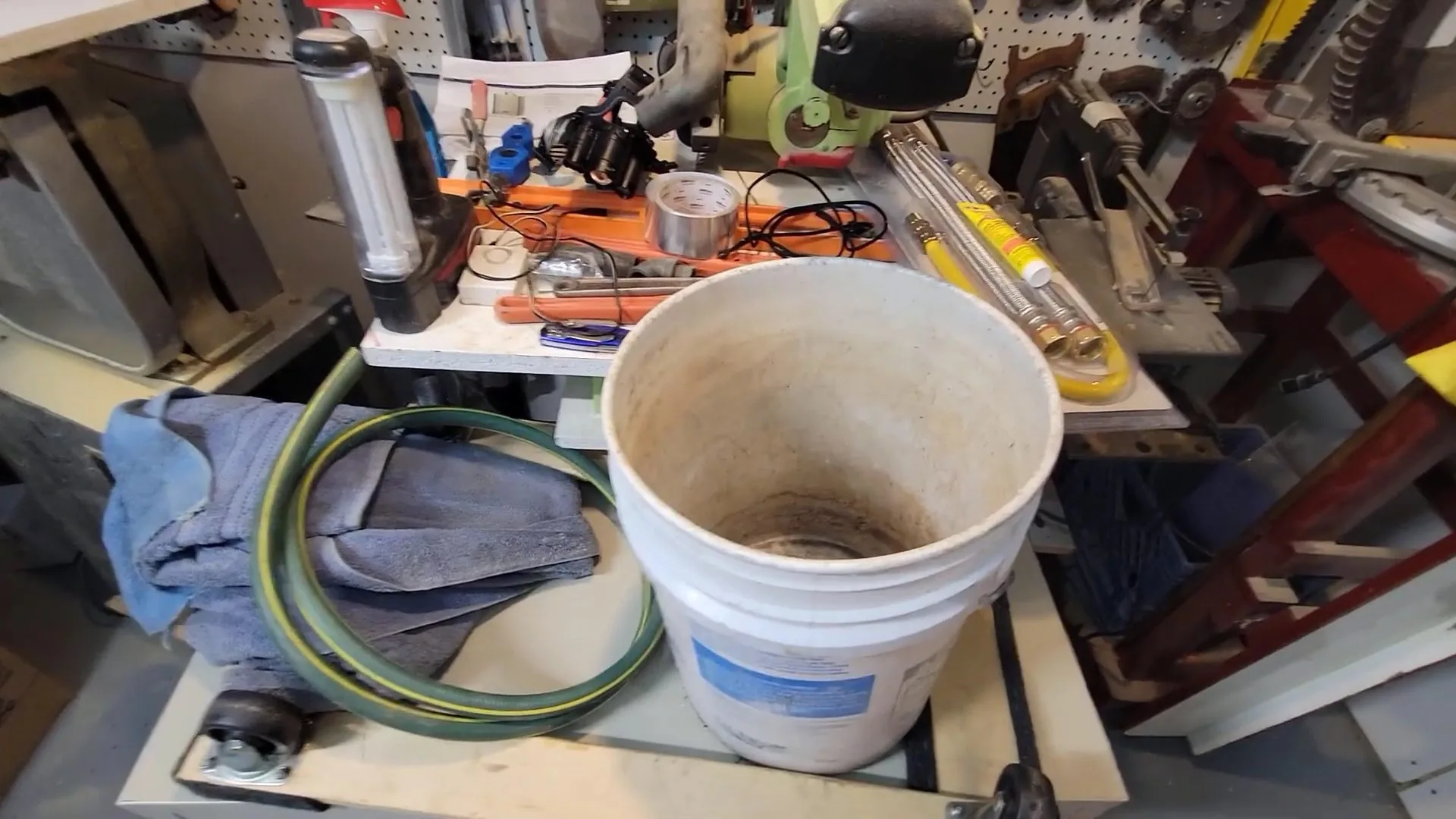 A plumber's workbench with a white bucket, a green hose, copper fittings and hand tools laid out