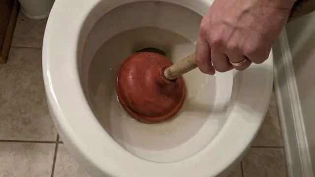 A hand pushing a red plunger down into a white toilet bowl