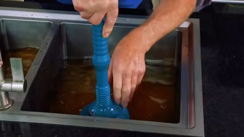 A hand working a blue plunger in a stainless sink that is holding brown standing water