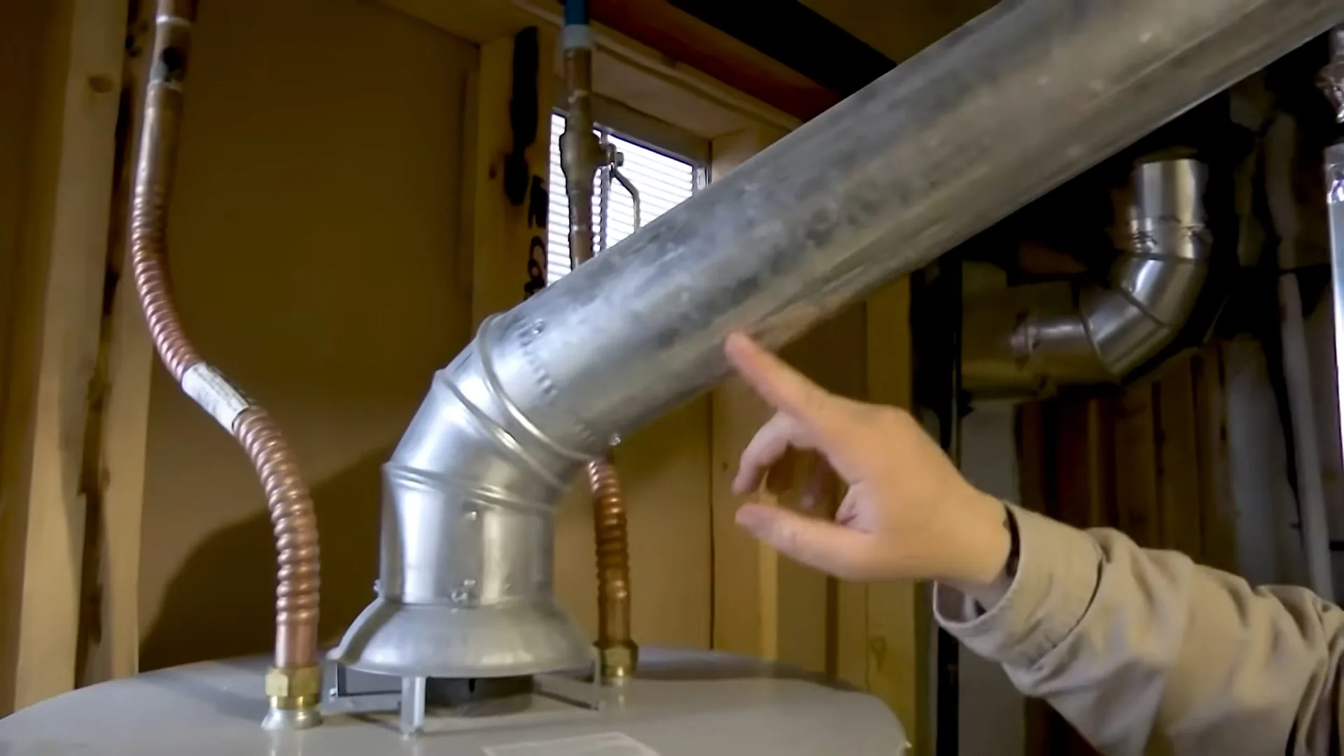 A hand pointing at a galvanized vent pipe rising from the top of a gas water heater between wooden studs