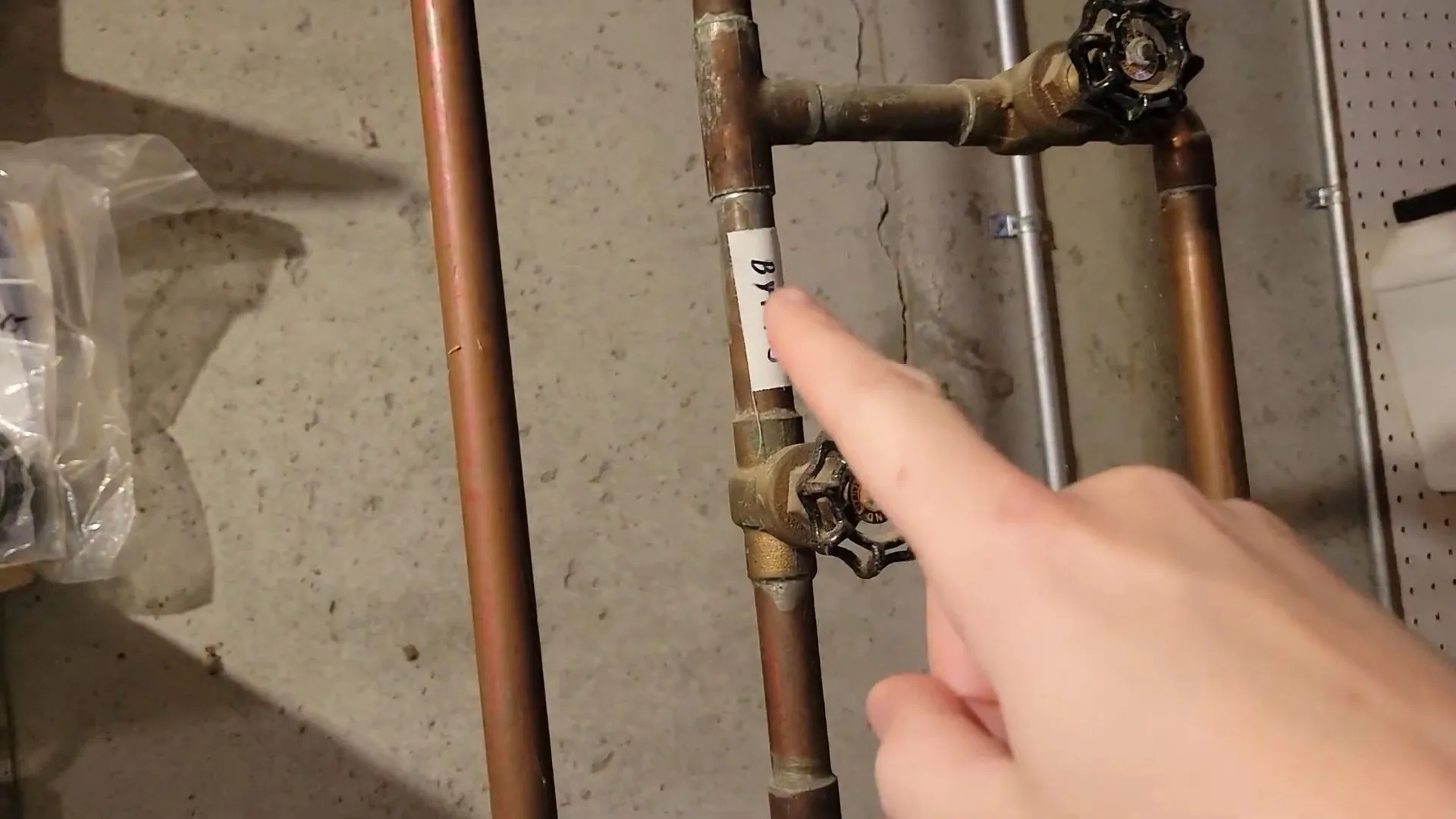 A finger pointing at a valve on a pair of copper supply pipes mounted on a basement wall