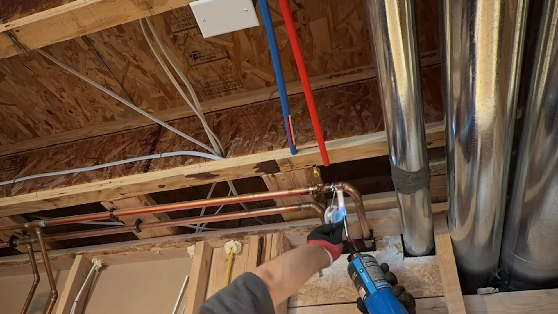 Red and blue PEX lines running between joists beside metal ducts, with a tool in a hand at the lower edge