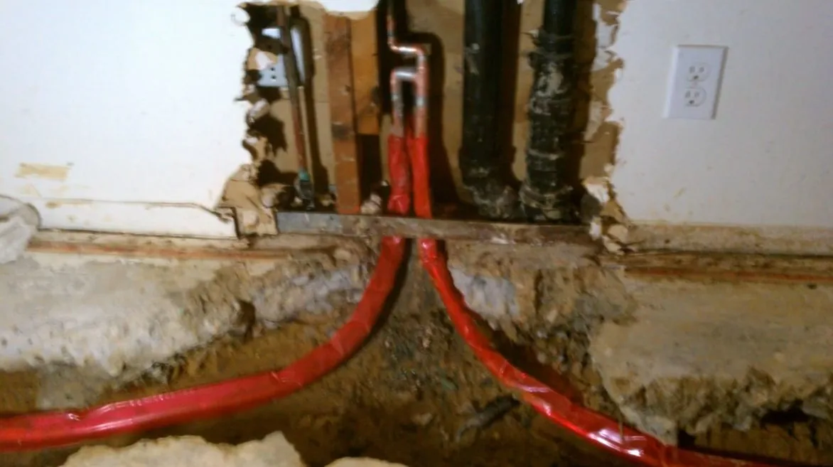 Red PEX supply lines running down through an opened wall and under the slab edge beside a black drain pipe