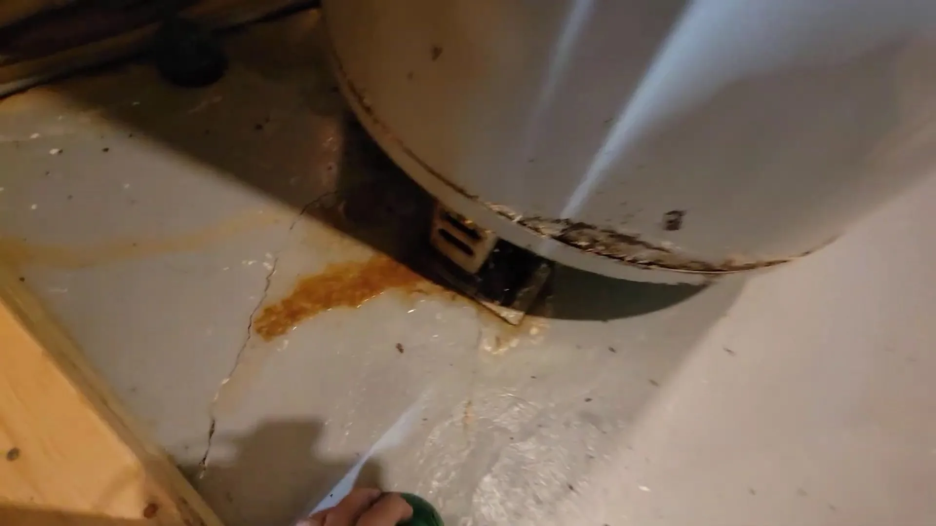 The lower edge of a water heater tank with a rusty orange stain on the floor beneath it