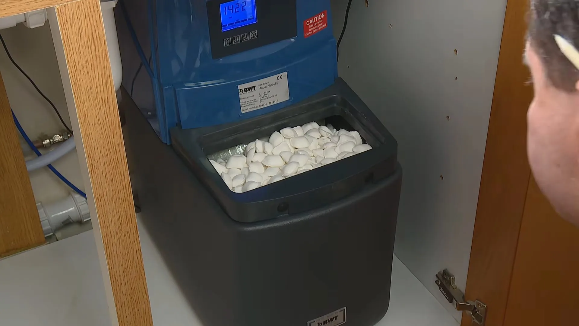 A water softener tucked in a cabinet with its lid off and white salt pellets filling the brine tank