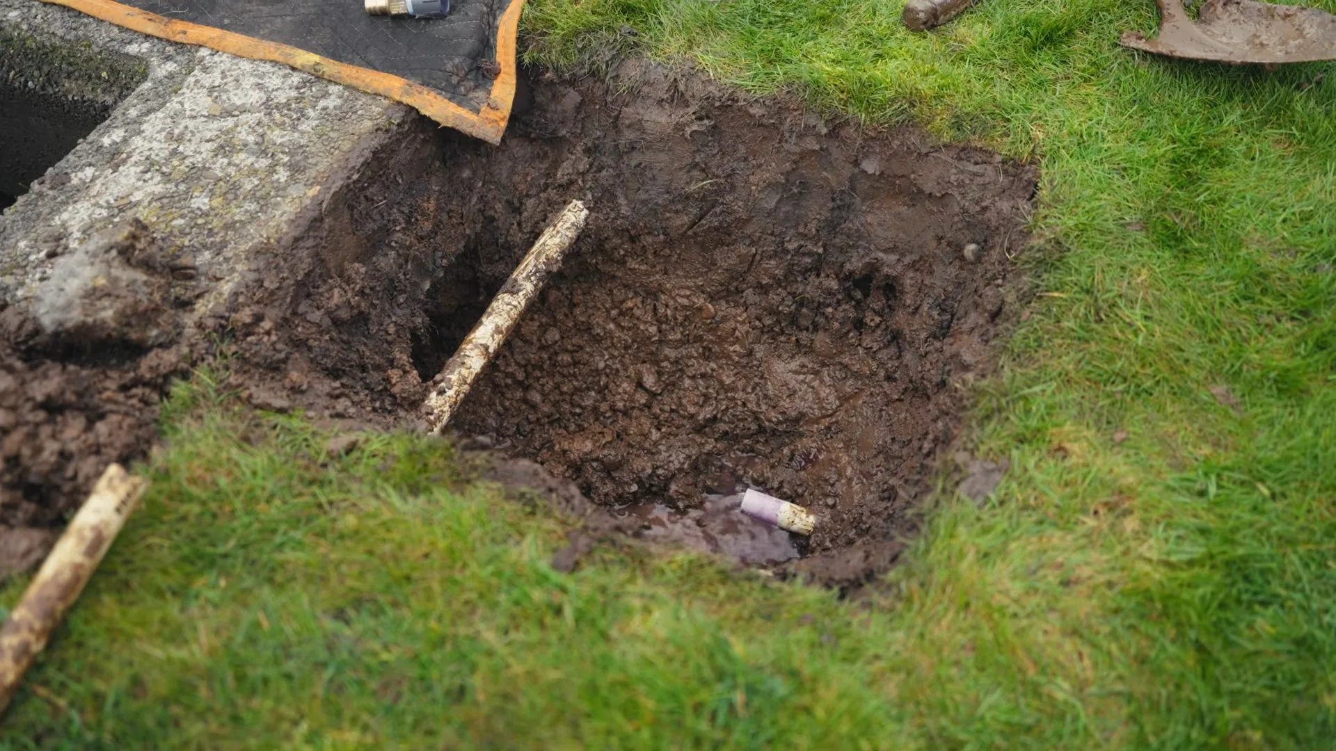 A hole dug in a lawn beside a concrete meter box, with wet soil, a wooden stick across the hole and a short length of pipe at the bottom