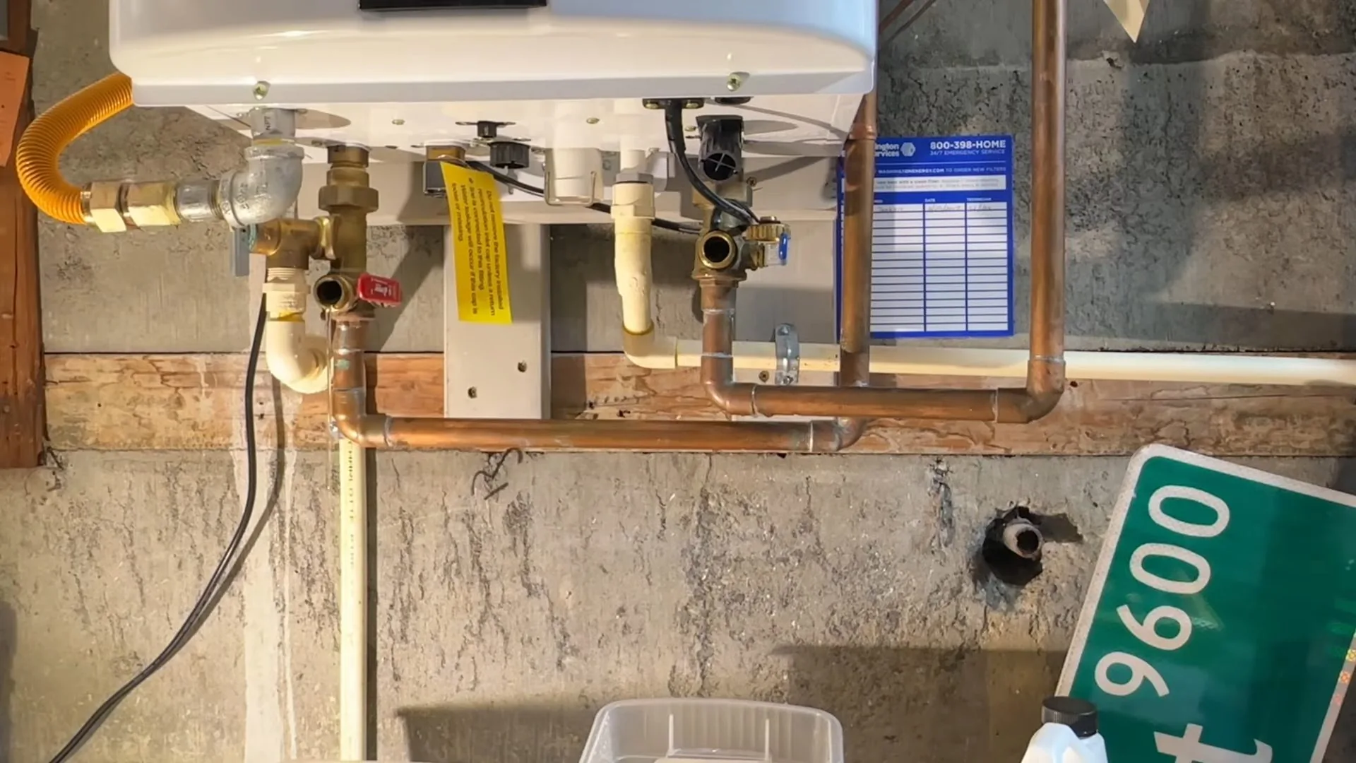 The underside of a tankless water heater with a yellow gas connector, copper pipes and brass valves above a concrete floor