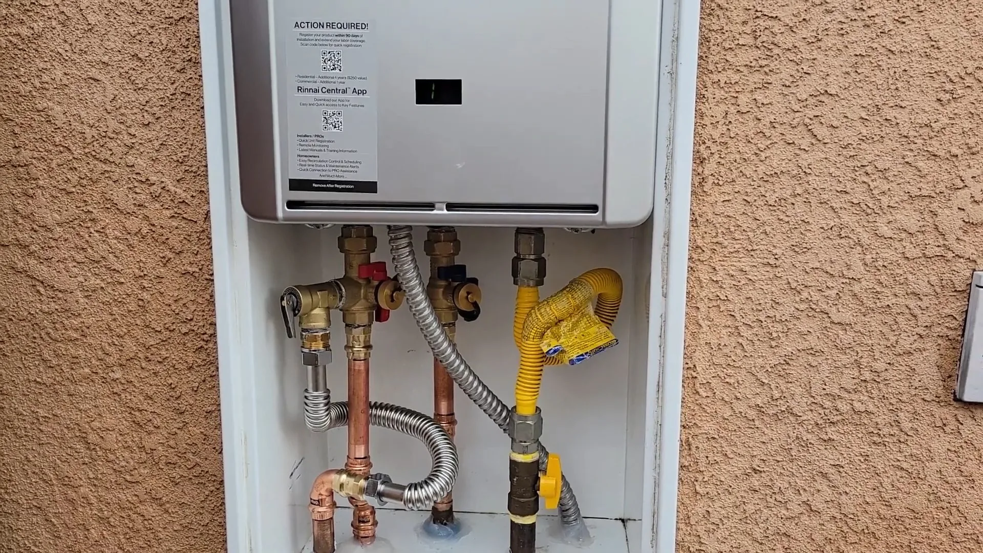 A wall-mounted tankless water heater in a recessed box on a stucco wall with gas and water connections underneath