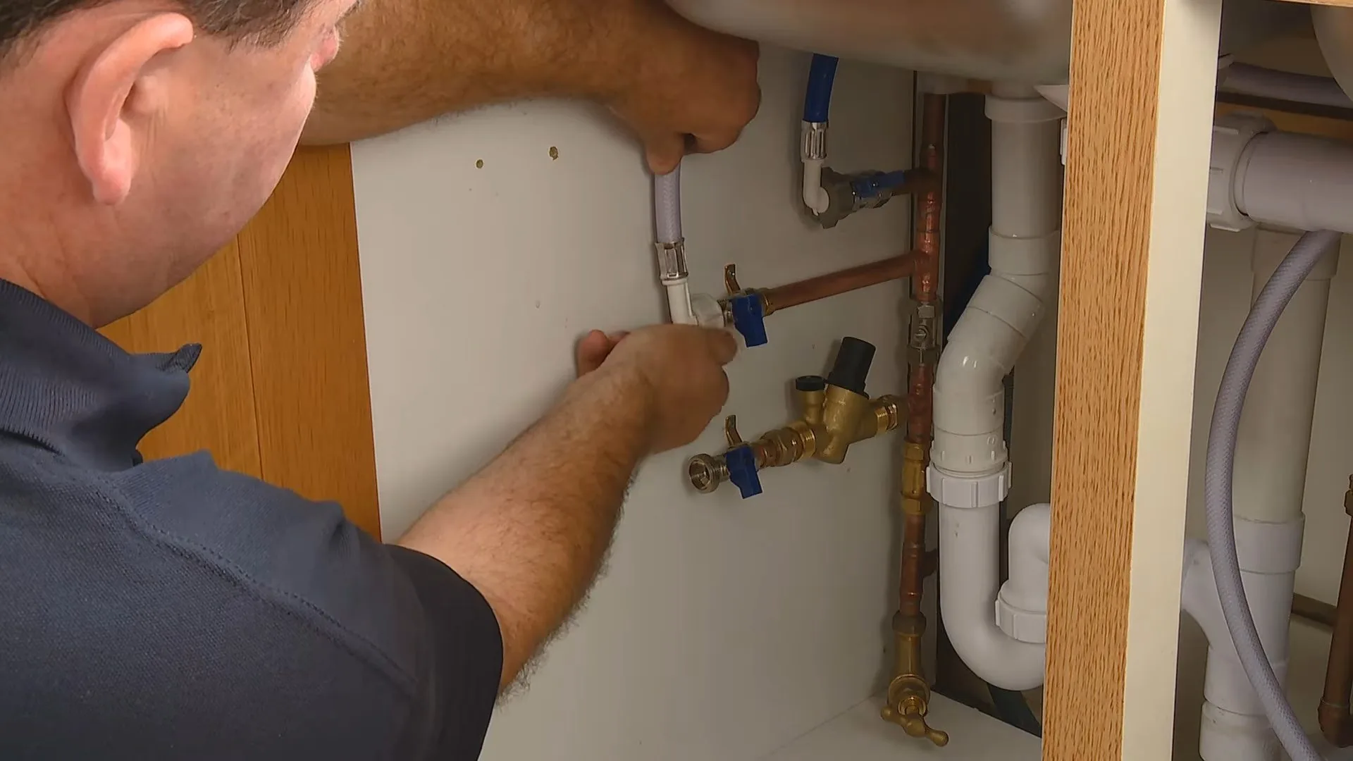 A technician reaching under a sink cabinet to connect a supply line at the shutoff valves, with the drain trap beside him