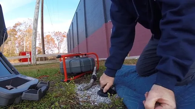 A technician kneeling on grass beside a cleanout behind a building, with a camera reel set out next to him