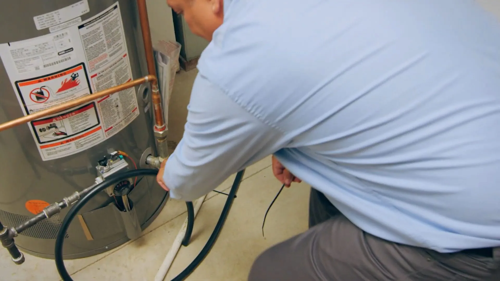 A technician in a light blue shirt kneeling beside a gas tank water heater with a hose connected at the base