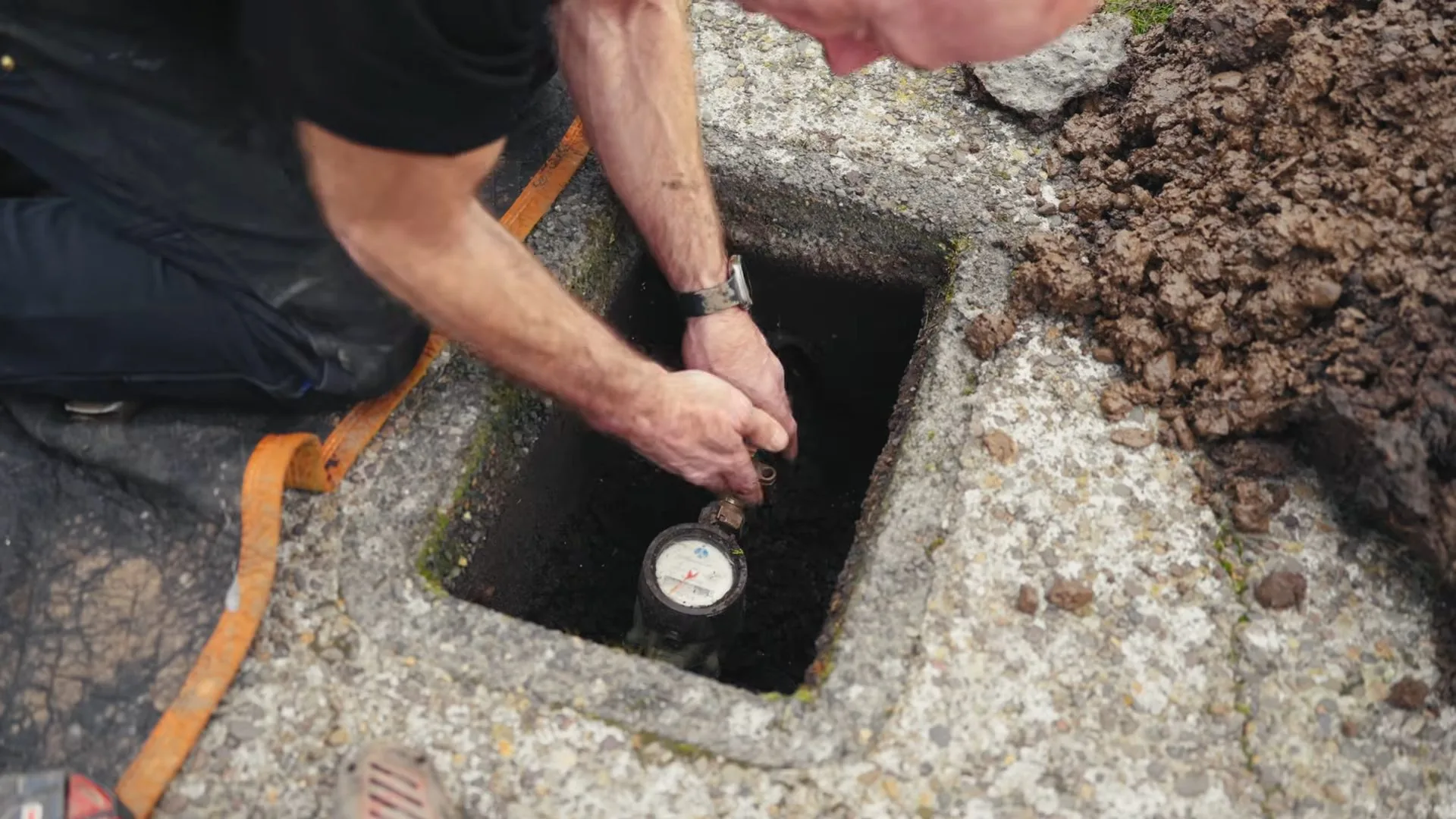A man leaning over an in-ground meter box beside the lid with his hand on the meter