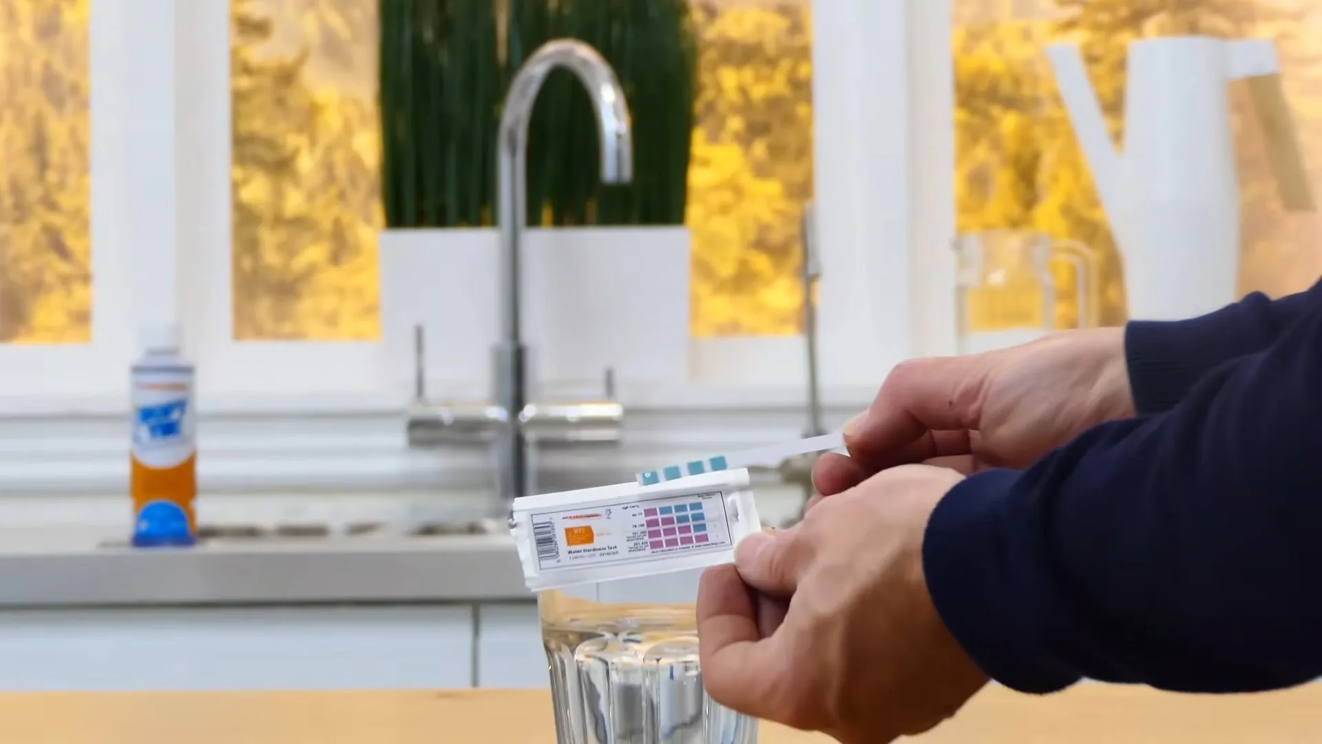 A hand dipping a test strip into a glass of water beside a kitchen faucet