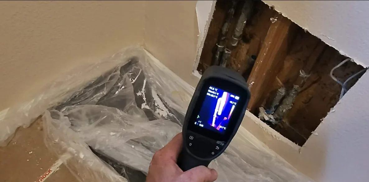 A hand holding a thermal camera whose screen shows bright and dark areas, pointed at an opened wall corner