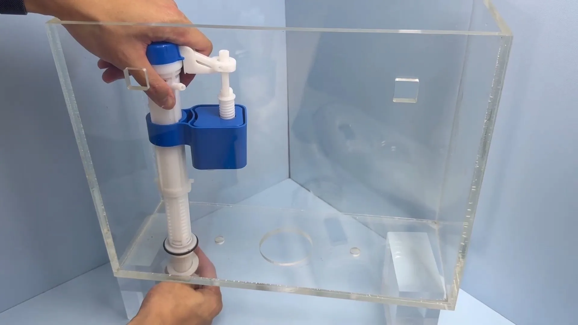 A hand holding the stem of a blue and white fill valve inside a clear acrylic demonstration toilet tank